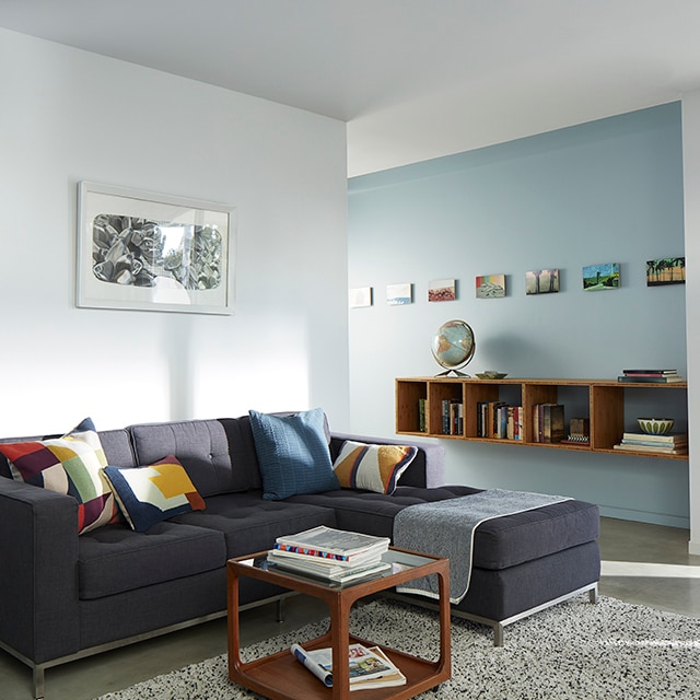 Sun cascading into a light blue and white painted living room with a dark gray sectional sofa and colorful pillows, open to a light blue hallway wall with a mounted bookcase and framed artwork.