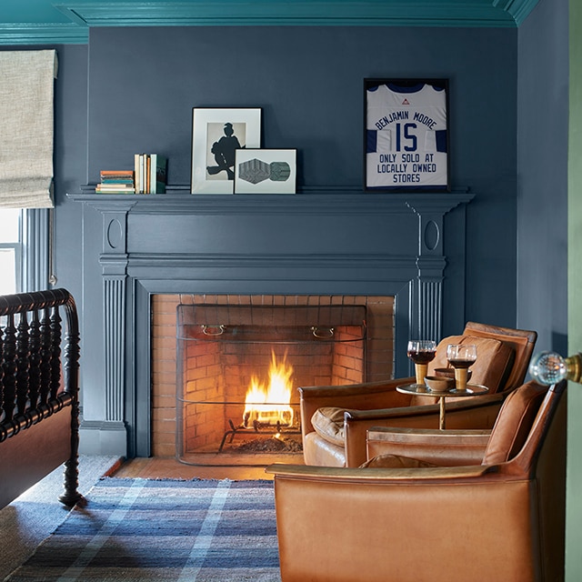 A cozy corner of a room with dark teal-blue painted walls and working fireplace mantel, a teal ceiling and crown molding, two brown leather chairs, a blue plaid rug, and the edge of a green door.
