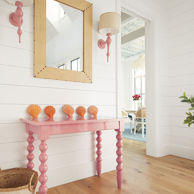 Una brillante entrada con paredes con revestimiento pintado de blanco, una mesa consola pintada de rosa claro, apliques de pared pintados de rosa y un espejo de ratán.