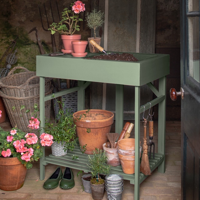Una caseta de jardinería pintada de verde musgo con macetas de arcilla, flores rosas, herramientas y cestas contribuye a crear un espacio de trabajo alegre dentro del cobertizo.