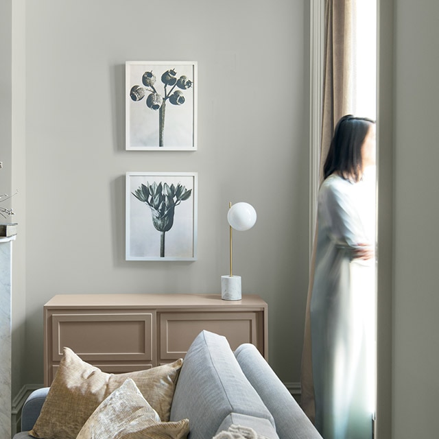 Una habitación acogedora en matices neutros y paredes pintadas de gris, un gabinete tostado elegante, un sofá gris y una mujer parada junto a una ventana.