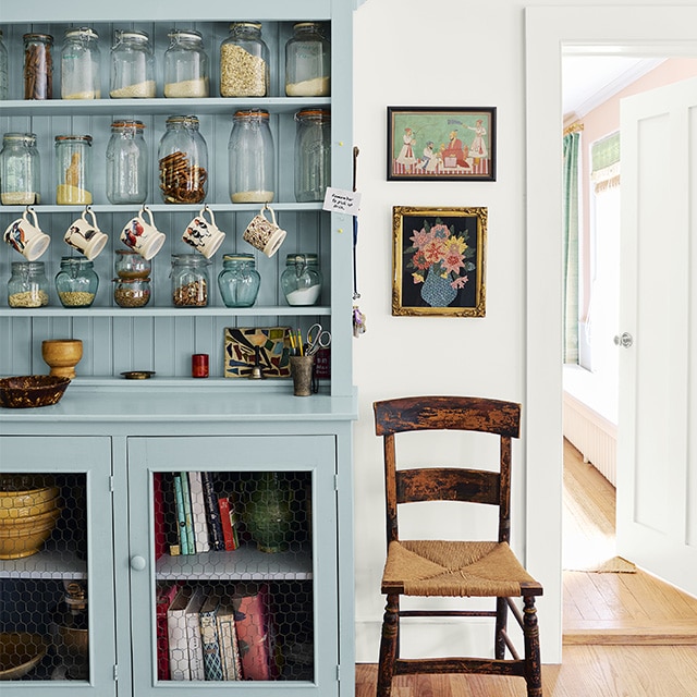 Joli bahut bleu clair avec pots en verre et livres à côté d’une chaise en paille posée contre un mur blanc près d’une porte ouvrant sur un mur arrière rose pâle.