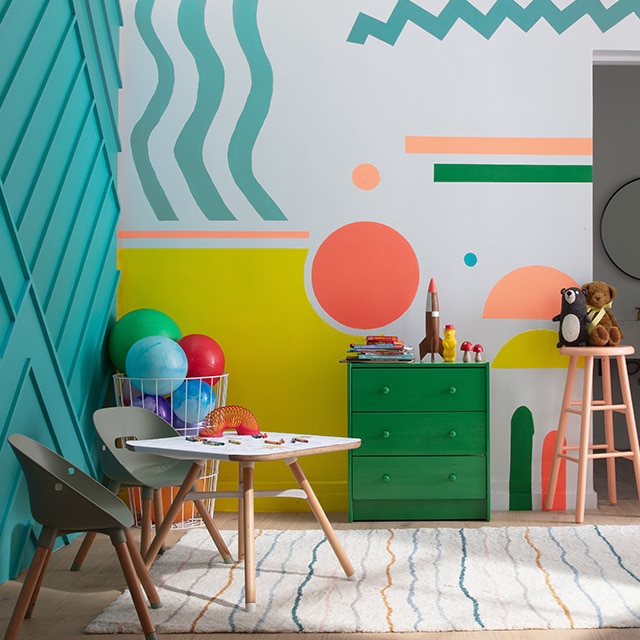 Une commode verte et une table et un tabouret pêche ajoutent à l’ambiance ludique de cette salle de jeux pour enfants avec un mur en planches avec couvre-joints turquoise et un autre mur blanc aux motifs géométriques jaune, pêche et turquoise.