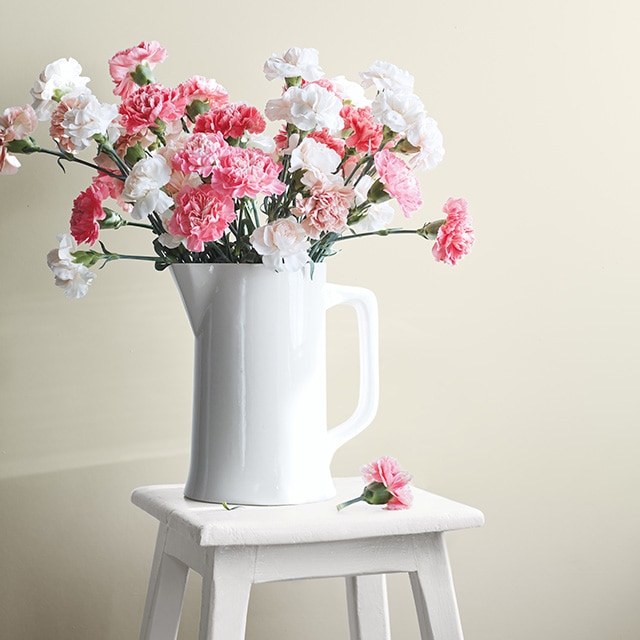 Un pichet blanc contenant des fleurs roses et blanches est posé sur un tabouret blanc devant un mur gris-vert pâle.