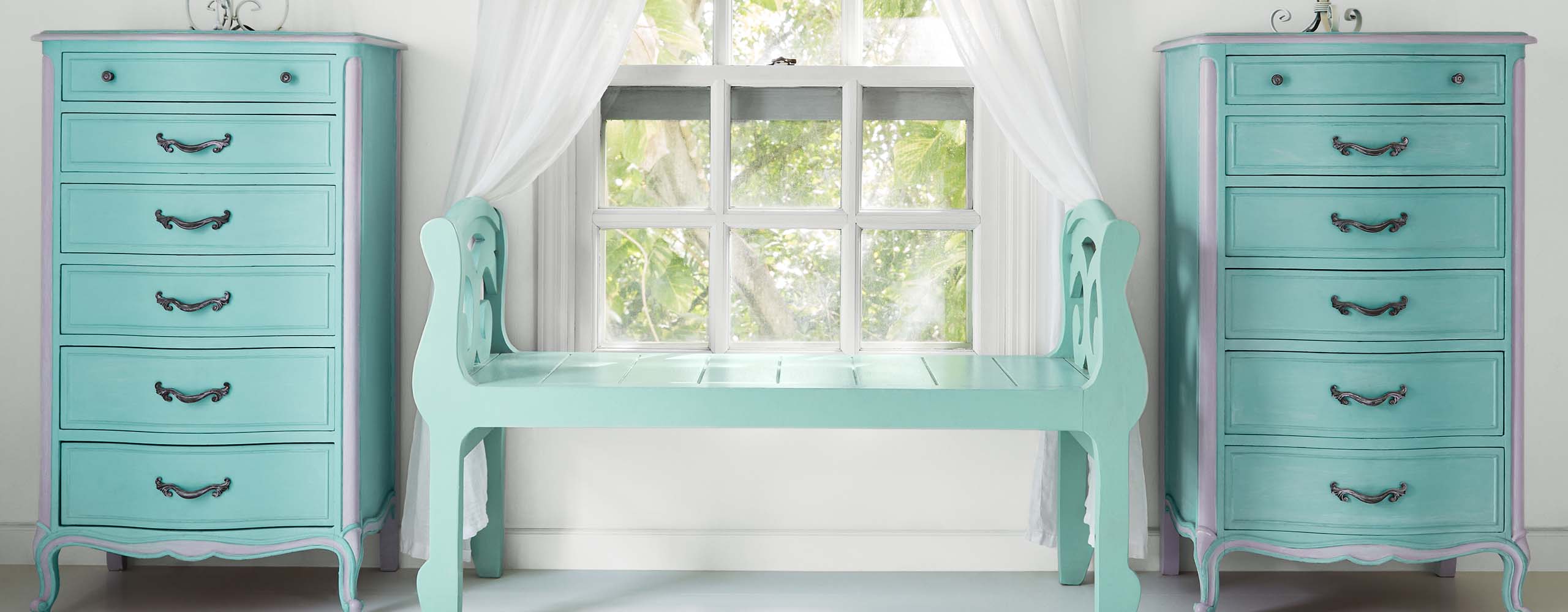 Une chambre à coucher joyeuse, peinte en blanc avec une touche de bleu-gris, un banc turquoise, une fenêtre avec des rideaux blancs flanquée de commodes peintes en bleu turquoise et une lampe sur chacune d’elles.