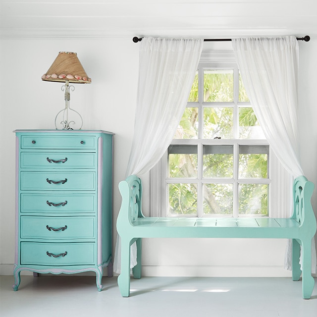 A cheery white-painted bedroom with a hint of blue-gray, a turquoise bench, and a window with white curtains flanked by turquoise-blue painted dressers with a lamp on each one.