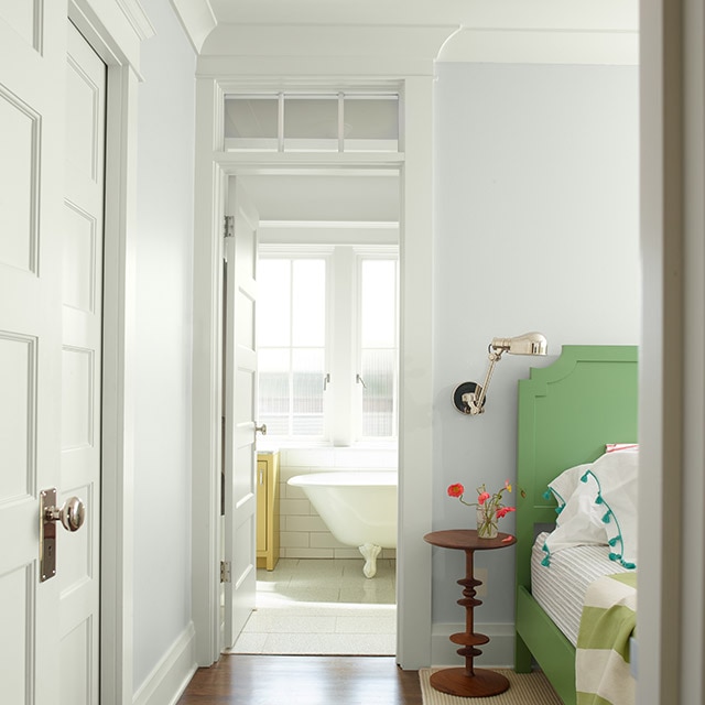 Una habitación acogedora pintada de blanco con cabecero verde, ropa de cama blanca y verde, una pequeña mesa auxiliar de madera y una puerta que se abre a un baño blanco con una bañera estilo antiguo.