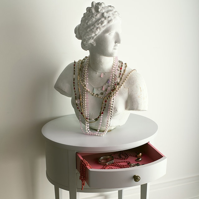 Un busto tallado con joyas colgadas en una mesa redonda pintada de gris con un cajón abierto color rojo escarlata en su interior frente a una pared pintada de blanco.