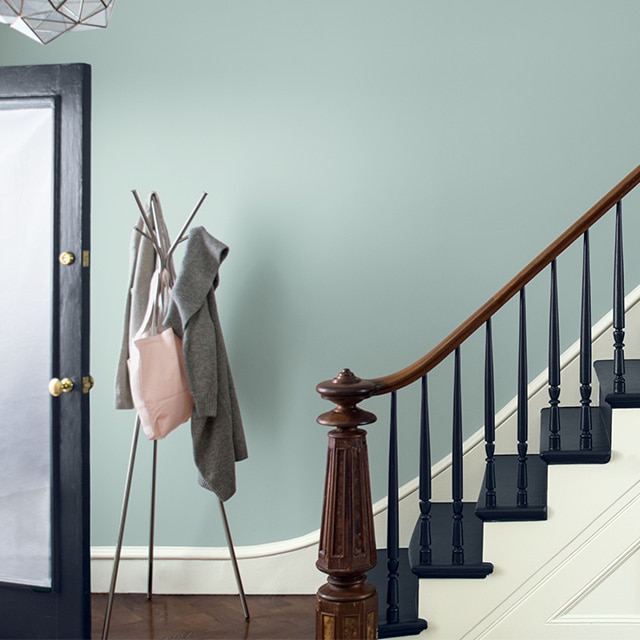 Una hermosa escalera de entrada pintada en azul marino con un pasamanos de madera y un poste central al lado de una pared pintada de gris azulado con marco blanco, y un perchero en la base de la escalera.