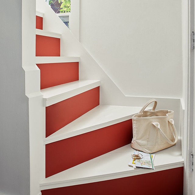 Una bella escalera con escalones pintados de blanco y contrahuellas en rojo brillante ofrece un gran contraste con las paredes pintadas de blanco y la ventana pequeña.