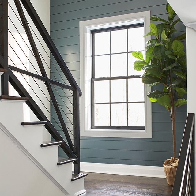 Una escalera de madera abierta y un rellano con contrahuellas pintadas de blanco y barandillas de metal negro, en una habitación pintada de blanco con una pared de acento traslapada y una ventana en gris azulado.