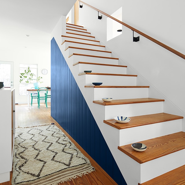 Una escalera pintada en blanco brillante con escalones de madera y una pared superior blanca, que dan hacia un pasillo con una pared en azul marino que conduce hacia una habitación trasera con sillas de color turquesa.