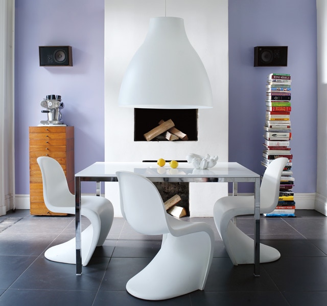 A white modern dining table and chairs in front of a pretty lilac painted wall with a white fireplace flanked by a tall stack of books and a tall set of drawers.