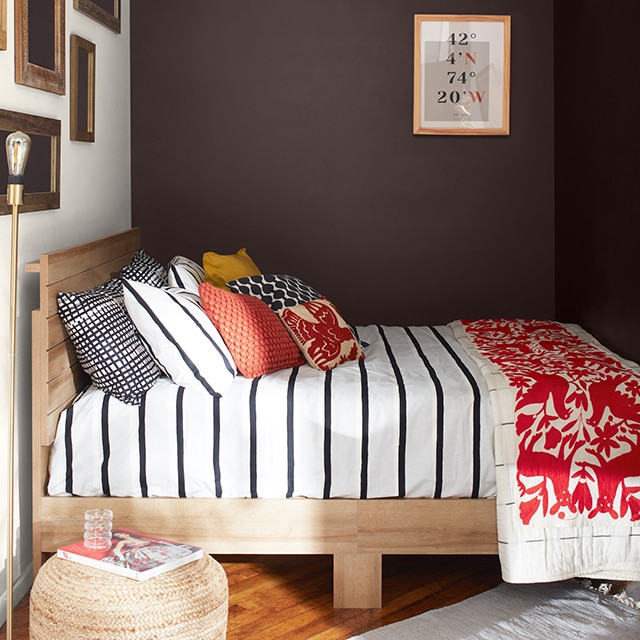 A cozy bedroom with deep chocolate-hued walls, a white-painted gallery wall, a bed with black and white striped bedding, pillows and red- printed blanket, and a wicker ottoman.