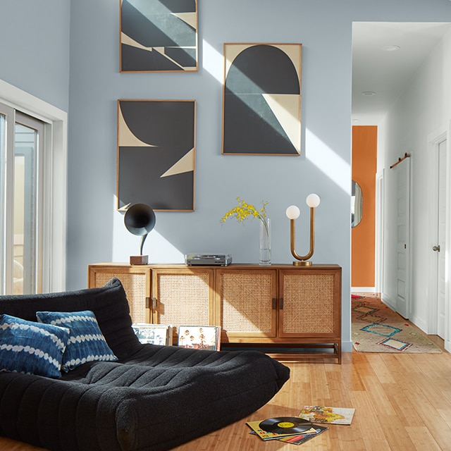 Una sala de estar luminosa con paredes pintadas de azul claro, techo blanco, un cómodo sofá oscuro, tres obras de arte moderno sobre un gabinete y una pared de acento naranja al final de un pasillo.