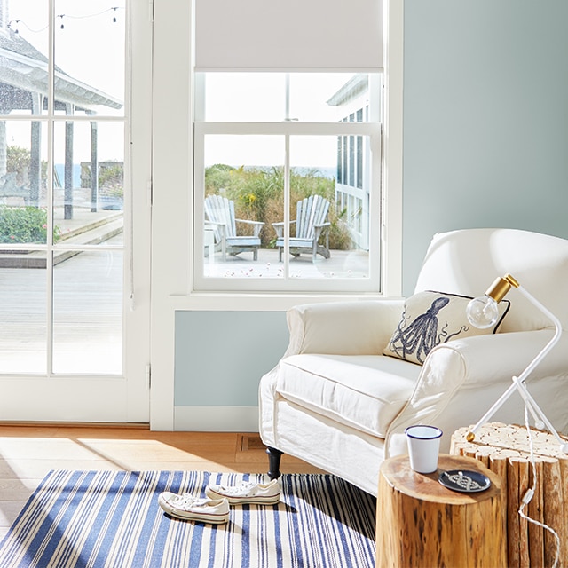 A sunny, light blue painted living room with a cozy white armchair, and an ocean view through a glass door and window.