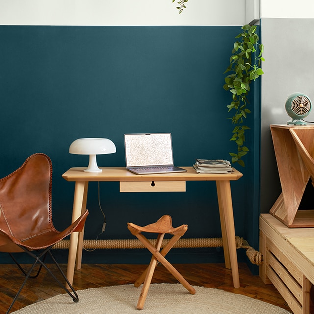A wood desk, leather butterfly chair, and stool in front of a two-tone white and teal blue painted wall with hanging plants on an upper shelf, and an adjacent white and gray two-tone wall.