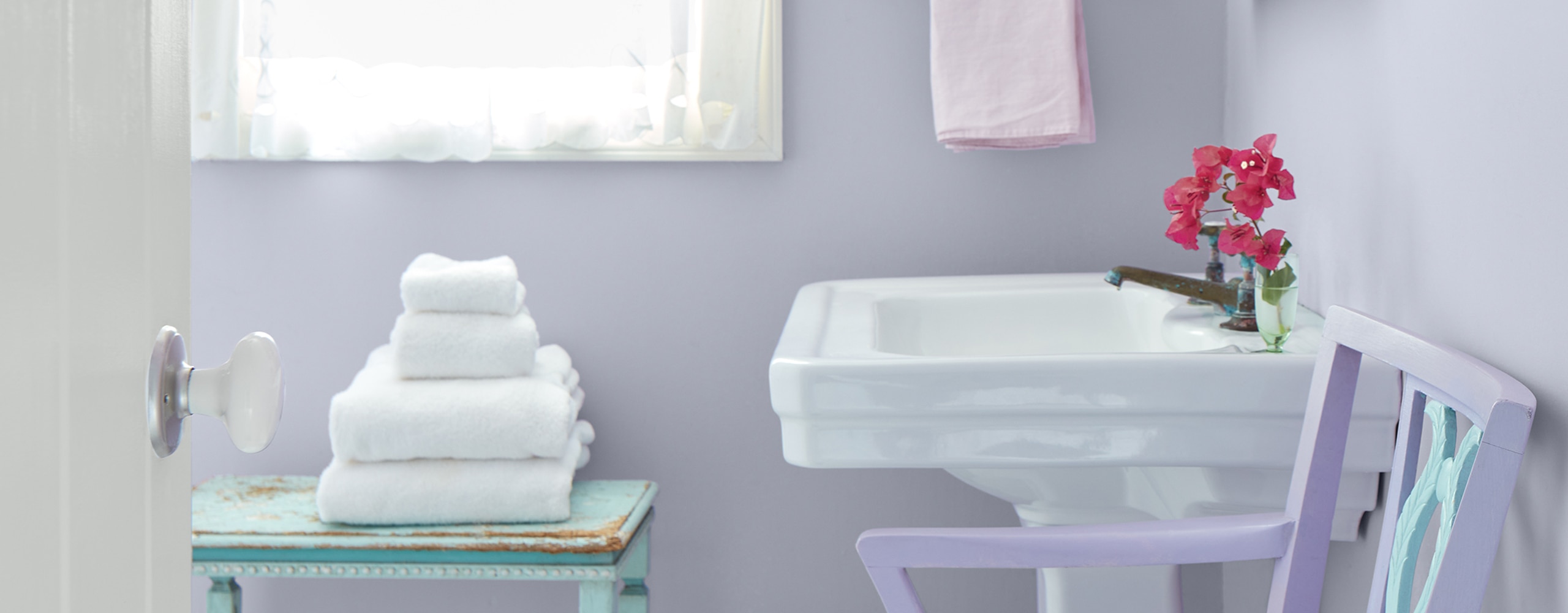 Un baño pequeño y soleado con paredes de color púrpura pastel, un lavabo blanco y una ventana con marco de blanco y cortinas blancas vaporosas.