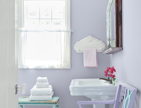 Un baño pequeño y soleado con paredes de color púrpura pastel, un lavabo blanco y una ventana con marco de blanco y cortinas blancas vaporosas.