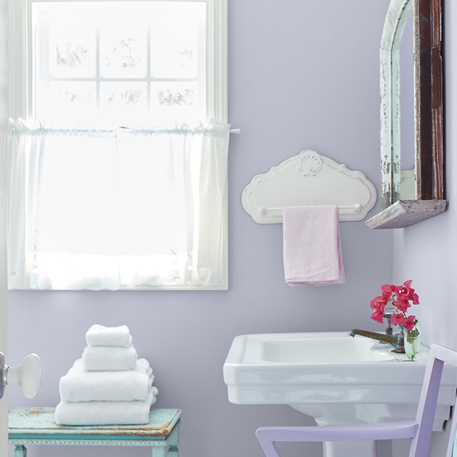 Salle de bains lumineuse aux murs violet pastel avec lavabo blanc, boiseries de fenêtre blanches et rideaux vaporeux blancs.