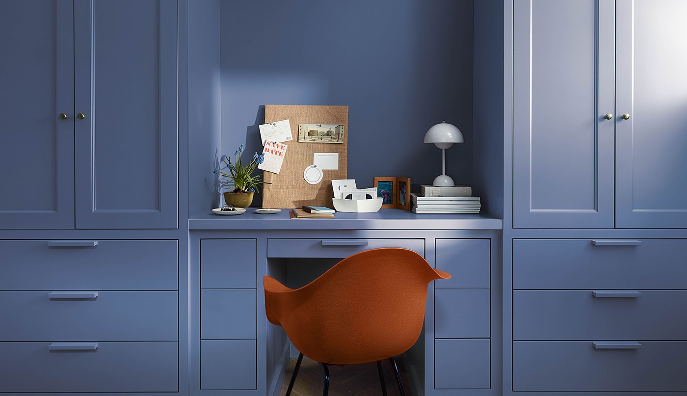 A beautiful home office space with blue painted built-in cabinets, desk, and drawers, and an orange chair.