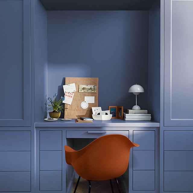 A beautiful home office space with blue painted built-in cabinets, desk, and drawers, and an orange chair.