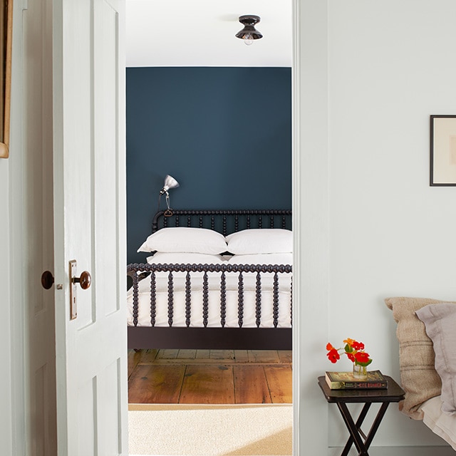 A white-painted room and white door open to a peaceful, dark blue bedroom with a white ceiling, white bedding, and a black spindle bed.