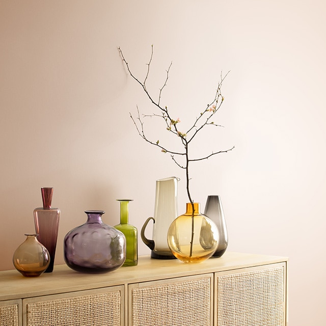 A dusty-pink painted wall behind a wood and rattan cabinet topped with colorful glass vases.