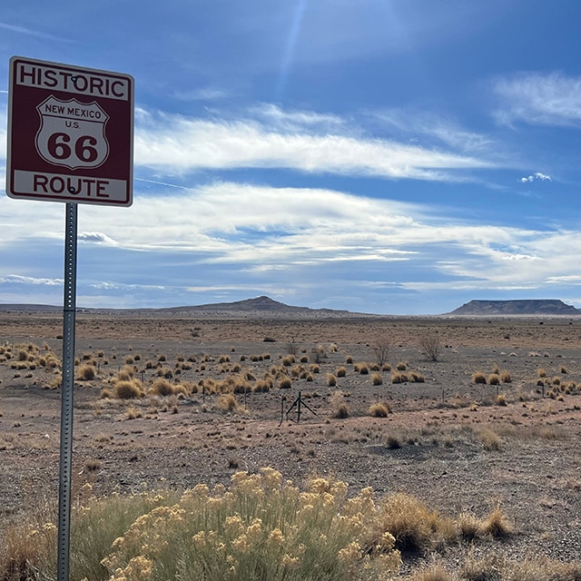 A historic Route Sixty-Six road sign stands alone in a desert landscape with a cloudy blue sky.
