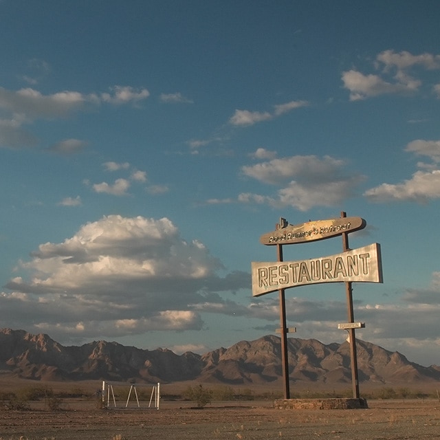 The Road Runner’s Retreat Sign sits along a dry desert highway with a cloudy blue sky and mountains in the distance.