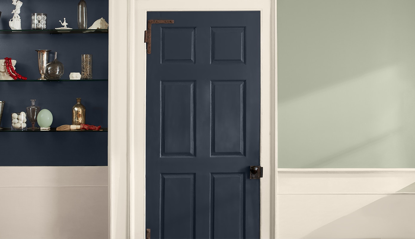 A two-tone wall painted light-sage green on the upper wall and off-white on the lower wall, with off-white trim, a dark blue door and inset shelves holding glass vases and trinkets.