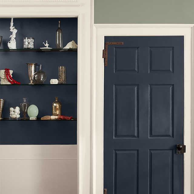 A two-tone wall painted light-sage green on the upper wall and off-white on the lower wall, with off-white trim, a dark blue door and inset shelves holding glass vases and trinkets.