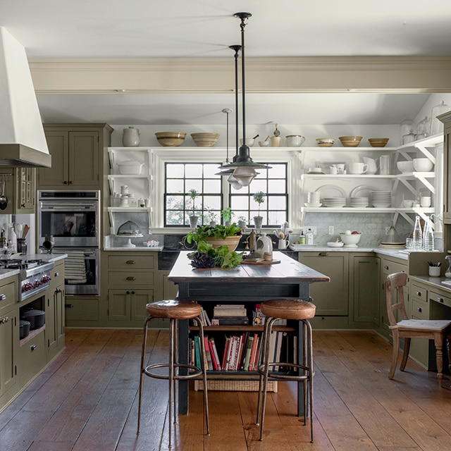 Una cocina abierta y acogedora, con gabinetes pintados de verde salvia, estantes abiertos blancos con una colección de cerámica de gres blanca, una isla central de madera rústica y piso de madera.