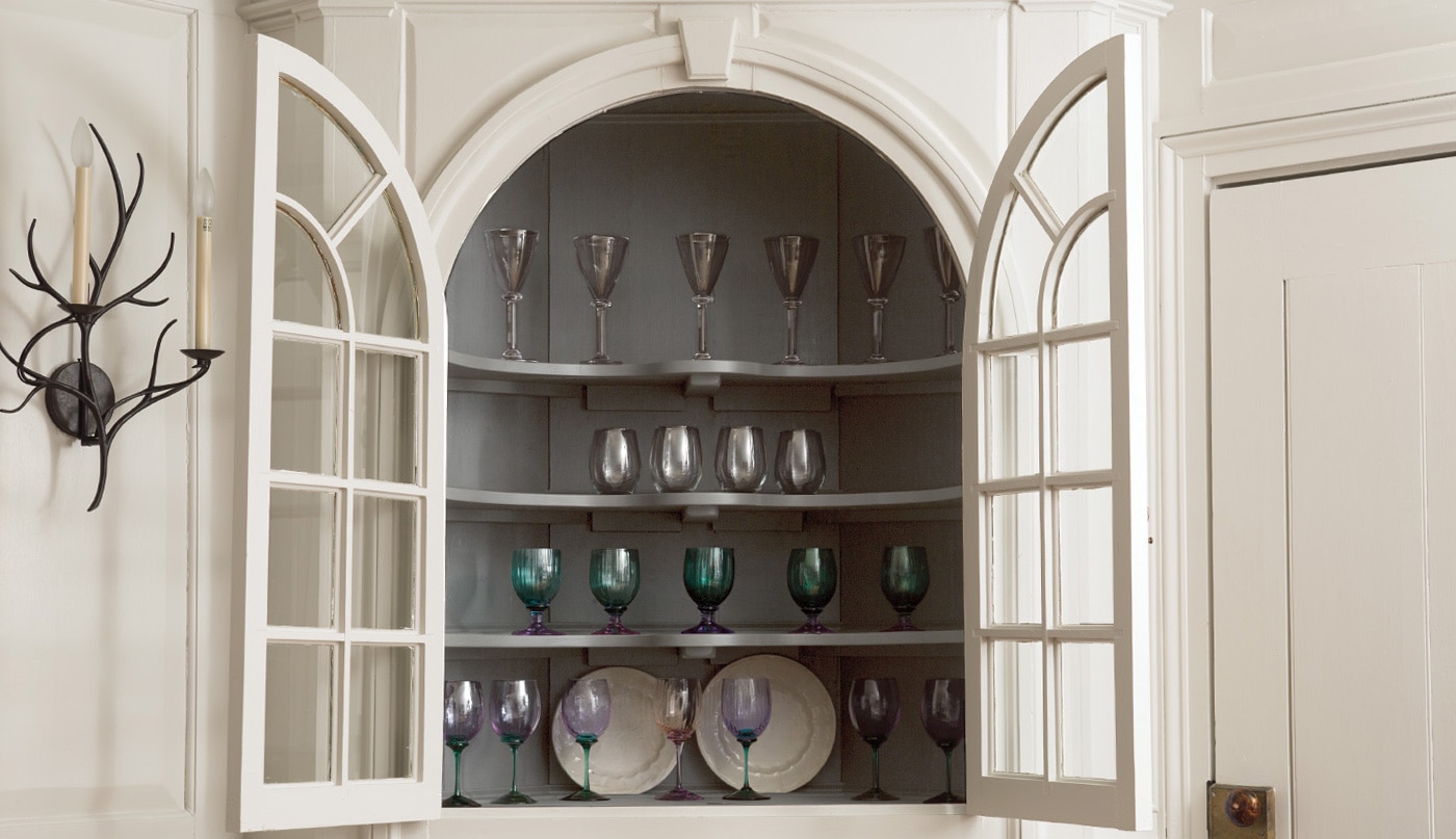A light-gray-painted built-in hutch with open glass doors, holding colored glassware and plates.