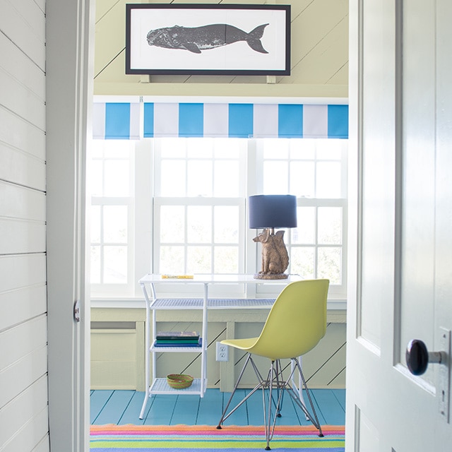 Couloir blanc avec porte blanche qui ouvre sur un bureau à l’ambiance de bord de mer avec mur et plafond en planches à feuillure jaune pâle, tableau de baleine suspendu au-dessus de plusieurs fenêtres, table blanche, chaise jaune et plancher bleu. 
