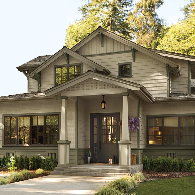 Una hermosa casa de estilo artesanal pintada de color Gris Pardo con techos a dos aguas y un porche en el frente, una puerta principal negra y detalles y marcos de color salvia, rodeada de vegetación.