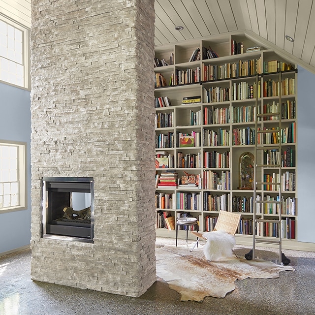 La biblioteca de la casa con estantes llamativos pintados de blanco desde el piso hasta el techo, paredes azules, marcos y techo abovedado con traslape blancos y chimenea central de piedra blanca.