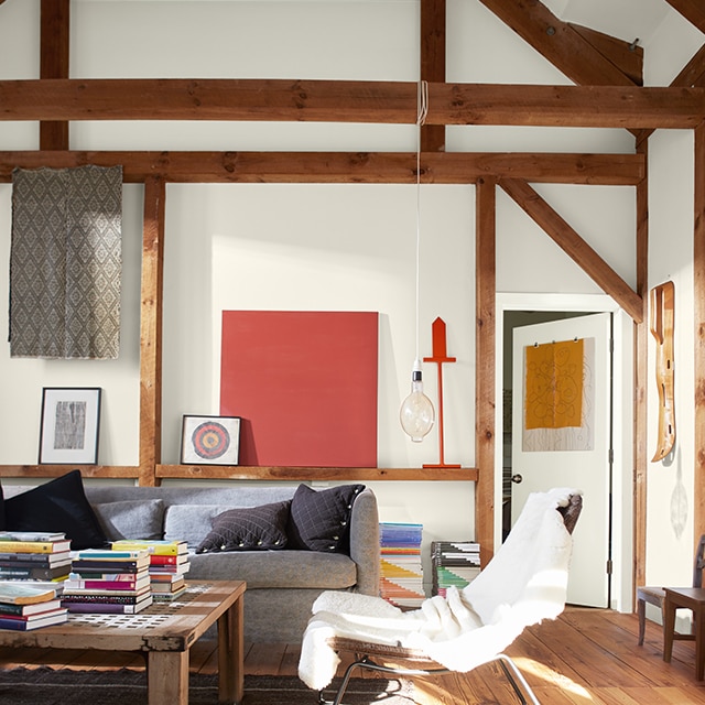 Una sala de estar pintada de blanco, iluminada por el sol, con techo abovedado y vigas de madera natural, arte en la pared, un sofá gris, una silla con una manta blanca y una mesa de café con libros.