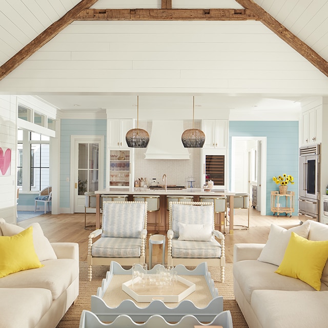 Una sala de estar y cocina aireadas pintadas de blanco con paredes con revestimiento y techo abovedado, vigas de madera, decoración azul y blanca, y paredes al fondo azules.