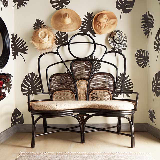 Paredes pintadas de blanco con plantillas con forma de hojas de palmera de color marrón oscuro, sombreros de paja colgados sobre un sillón para dos personas de mimbre y tela, y una alfombra beige.