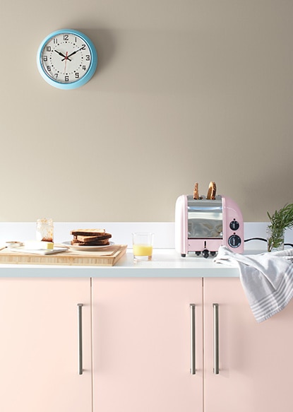 Une cuisine arborant des armoires peintes en rose pâle, un grille-pain rose, une planche à découper, de la vaisselle et un mur peint en brun clair avec une horloge bleue.