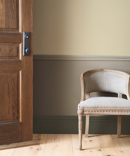 An open wood door lets light in to a hallway featuring an antique accent chair set against earthy green-toned wainscotting.