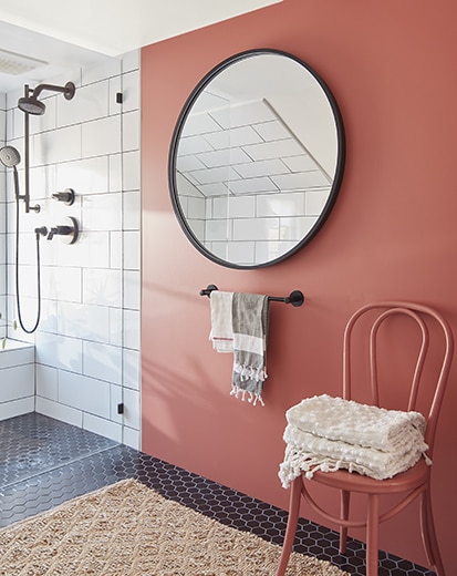 Salle de bains rouge terre avec douche en carreaux blancs, miroir rond, chaise avec serviettes, porte-serviettes et petit tapis beige.