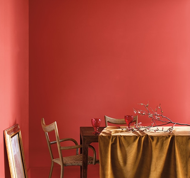 Red orange dining room painted in Raspberry Blush 2008-30 with wooden table, gold tablecloth, tree branches, and a golden frame propped against the wall.