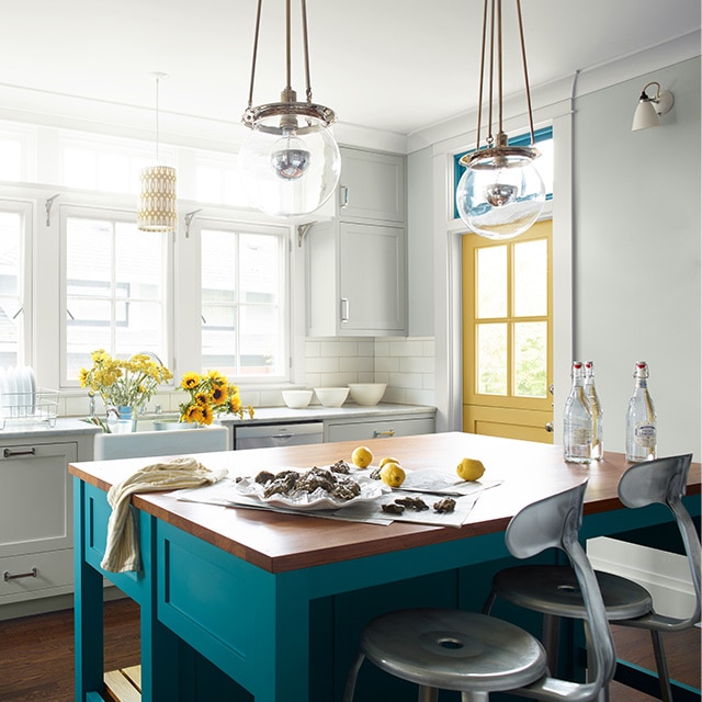 White kitchen with blue-painted kitchen island, yellow door, modern light fixtures, and large windows.