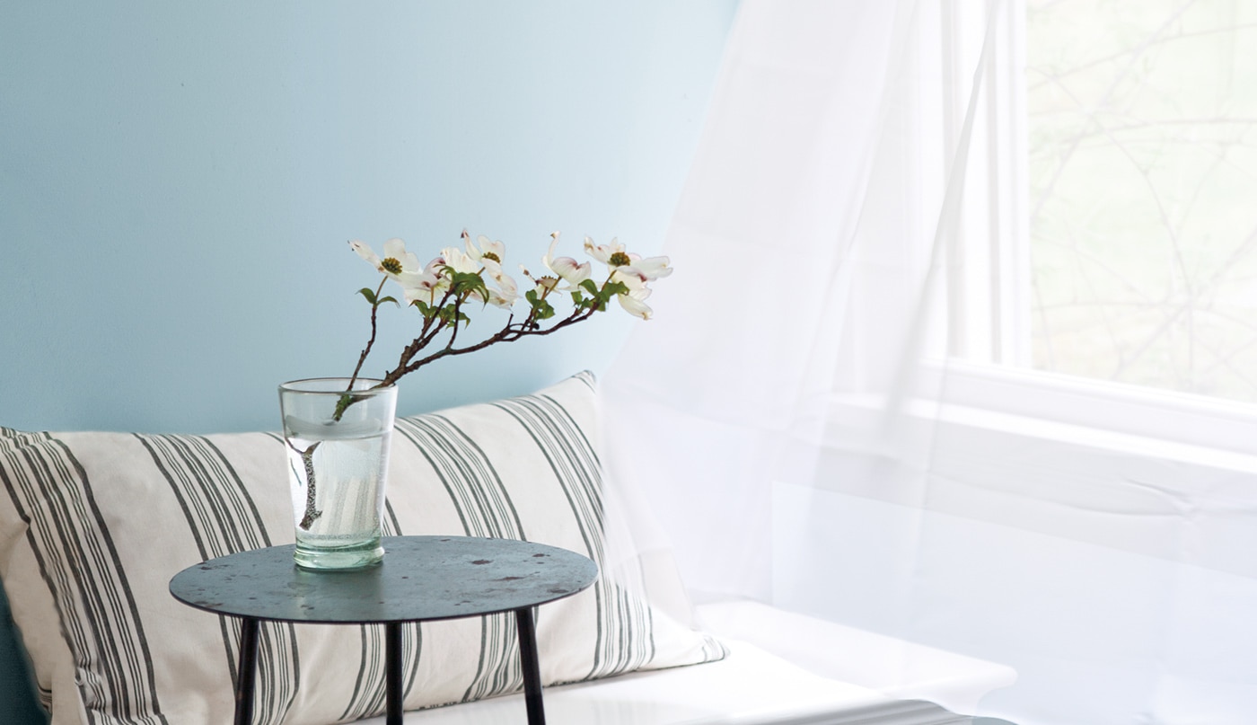 Un mur peint en bleu clair, une fenêtre ouverte avec un voilage blanc flottant au vent, un banc orné d’un coussin rayé et une table d’appoint noire.