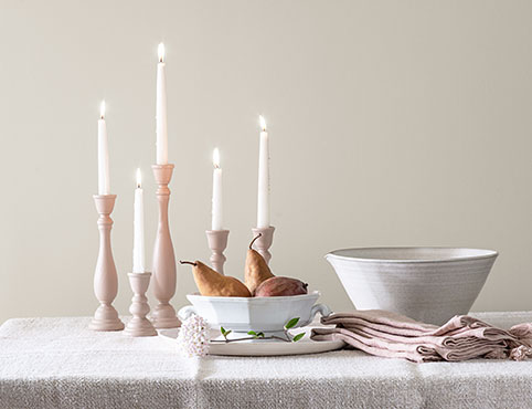 Lit tapered candles, 2 white porcelain bowls and off-white cloth napkins sit on a table against a white-painted wall.