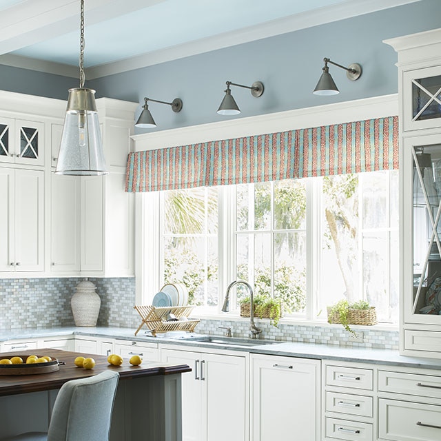 Cuisine accueillante aux murs bleu pâle avec plafond bleu glacé et poutres blanches, trois grandes fenêtres devant un évier, îlot de cuisine et armoires blanches.