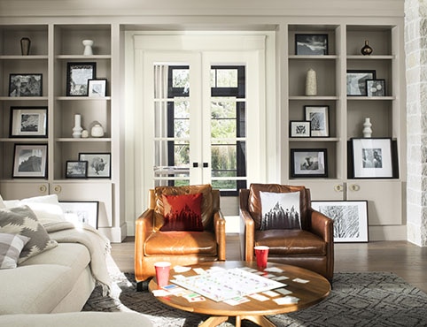 Salon confortable avec bibliothèques grises encastrées, portes-fenêtres blanches, sofa blanc et fauteuils en cuir brun.
