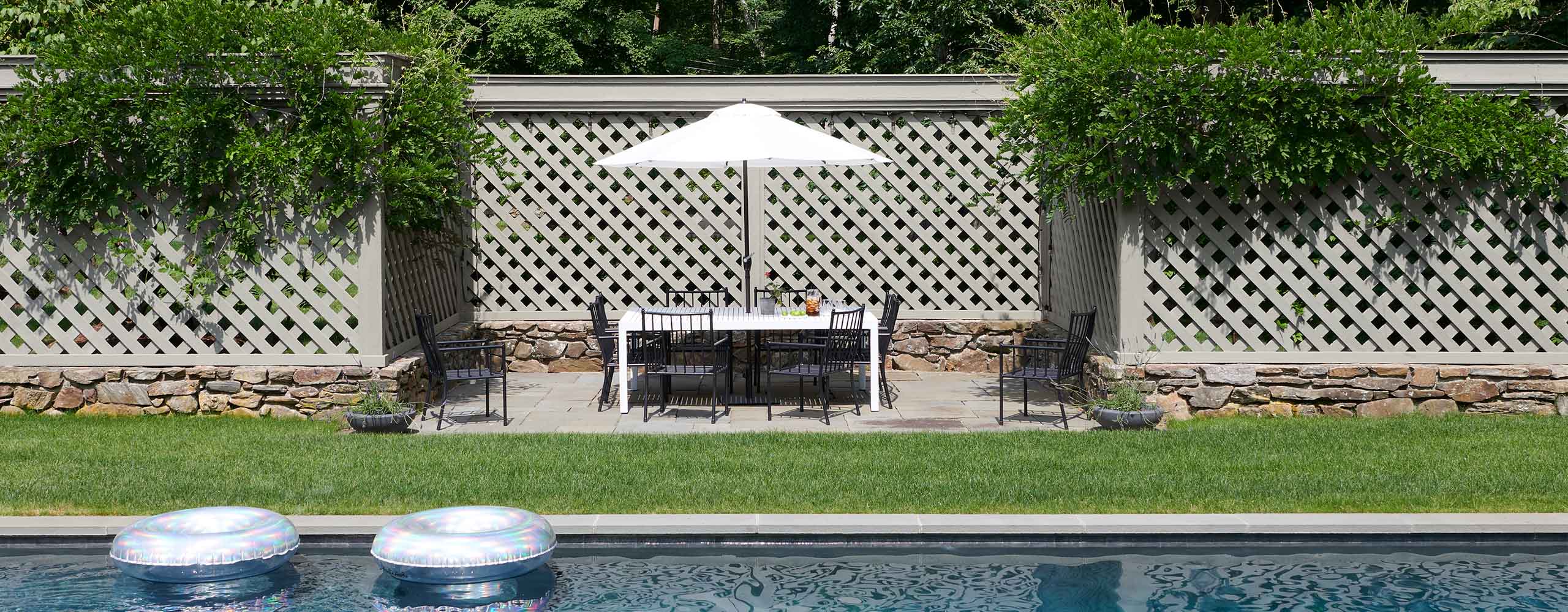 A beautiful gray-painted trellis style fence with climbing greenery offers privacy in this yard with a pool and outdoor patio with dining furniture and a white umbrella.